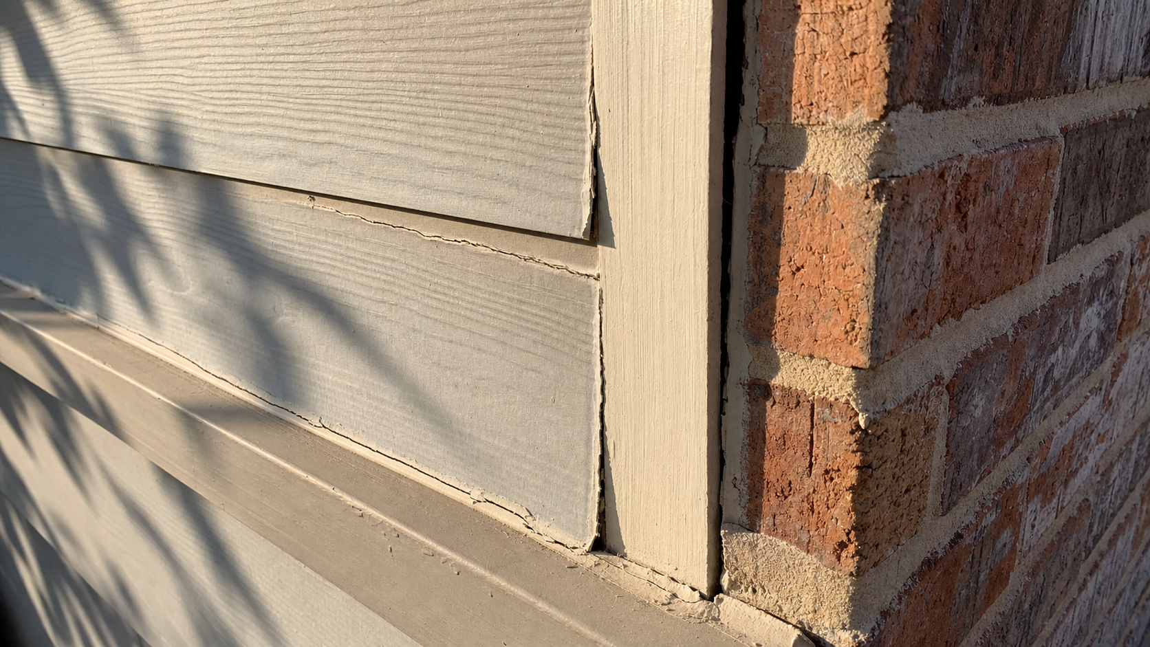 Sun-baked hardie siding corner meeting brick showing hairline caulk cracks on a Houston home exterior