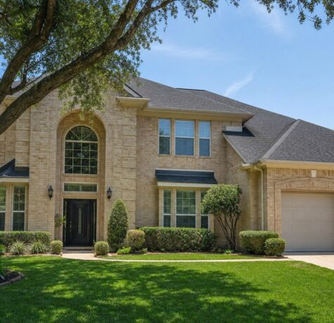 Two-story tan brick Houston home with new charcoal architectural shingle roof under clear midday sky