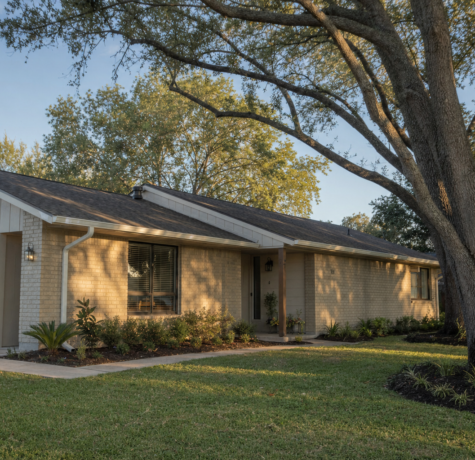 Houston home exterior in late September with mature oak canopy and visible shingle roof line